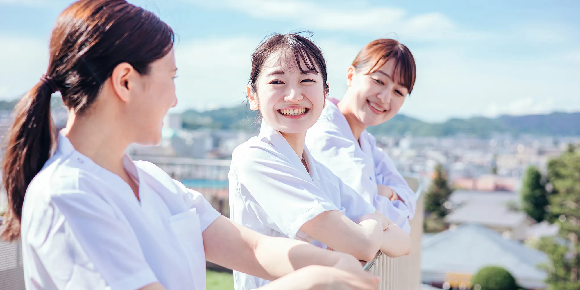 青空の下、テラスで笑顔で並ぶ3人の医療従事者（採用イメージ）